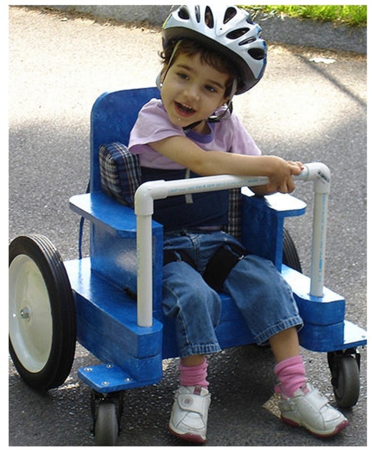 A child rides an adaptive wheeled riding toy, made of cardboard and PVC pipe.